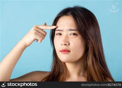 Beauty skin care concept. Asian young woman pointing eyebrow with her finger studio shot isolated on blue background, beautiful portrait female point face and forehead
