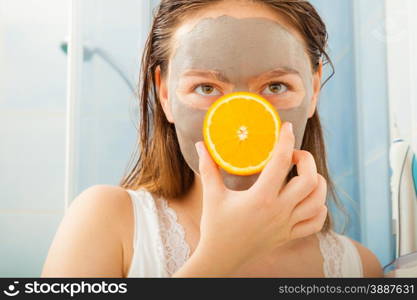 Beauty skin care cosmetics and health concept. Young woman in bathroom with facial clay mask holding orange fruit slice playing