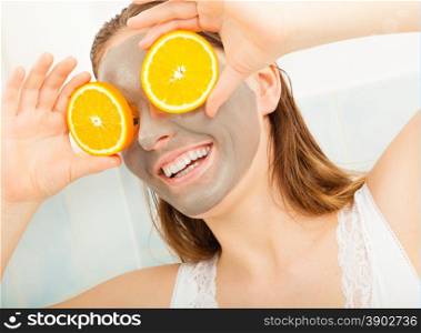 Beauty skin care cosmetics and health concept. Young woman with facial clay mask holding orange fruit slice covering eyes in bathroom