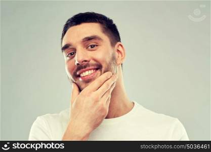 beauty, skin care, grooming and people concept - happy young man touching his face or beard over gray background. happy young man touching his face or beard