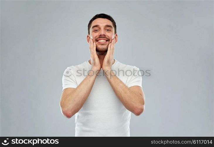 beauty, skin care, grooming and people concept - happy young man touching his face applying aftershave over gray background