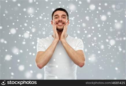 beauty, skin care, grooming, winter and people concept - happy young man touching his face applying aftershave over snow on gray background
