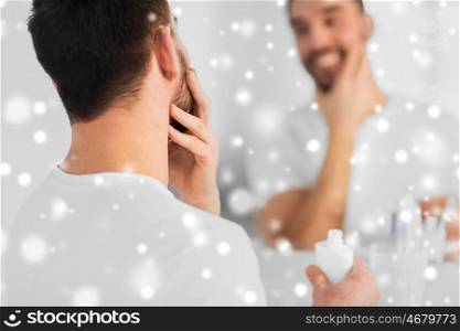 beauty, skin care, shaving and people concept - close up of smiling young man applying cream to face and looking to mirror at home bathroom over snow