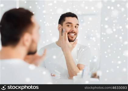beauty, skin care, winter and people concept - smiling young man applying cream to face and looking to mirror at home bathroom over snow
