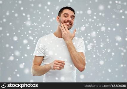 beauty, skin care, winter, christmas and people concept - smiling young man applying cream or lotion to face over snow on gray background