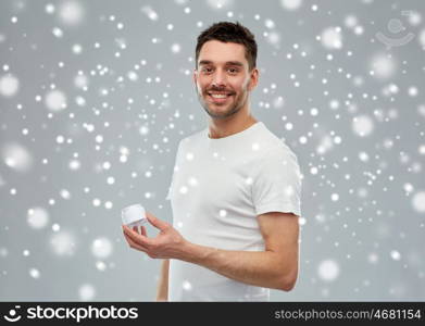 beauty, skin care, winter, christmas and people concept - smiling young man with cream jar over snow on gray background