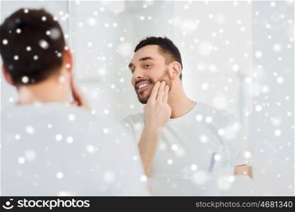 beauty, skin care, winter, christmas and people concept - smiling young man applying cream to face and looking to mirror at home bathroom over snow
