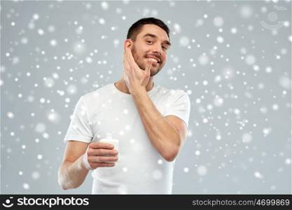 beauty, skin care, winter, christmas and people concept - smiling young man applying cream or lotion to face over snow on gray background