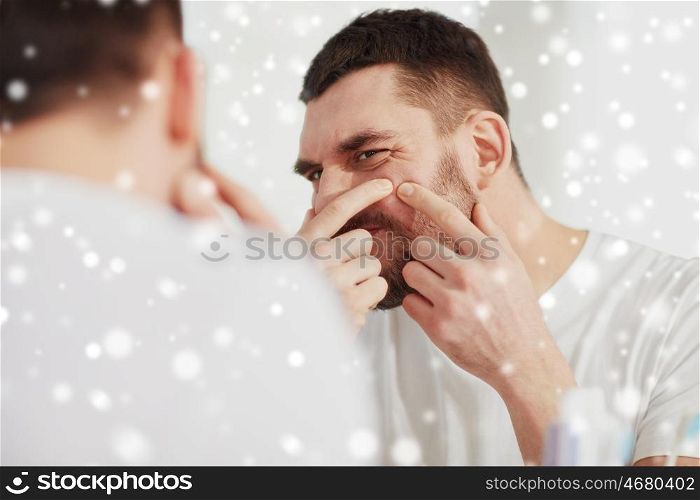 beauty, skin problem, winter, christmas and people concept - young man looking to mirror and squeezing pimple at home bathroom over snow