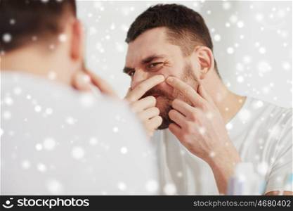 beauty, skin problem, winter, christmas and people concept - young man looking to mirror and squeezing pimple at home bathroom over snow