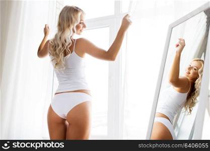 beauty, slimming and people concept - beautiful young woman in white underwear at mirror near window in morning at home. woman in underwear at mirror in morning