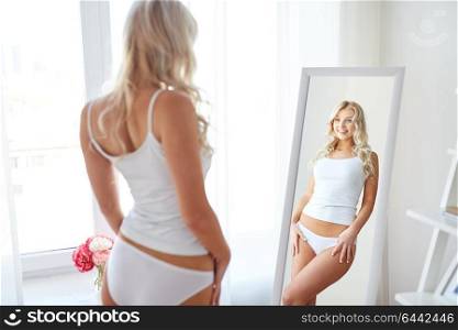 beauty, slimming and people concept - beautiful young woman in white underwear looking at mirror in morning at home. woman in underwear looking at mirror in morning
