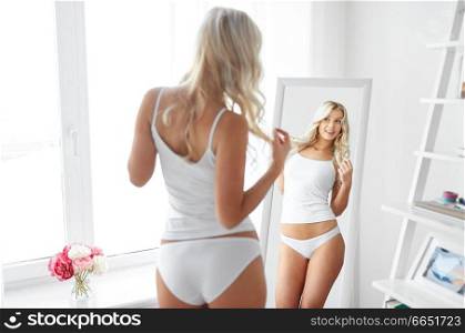 beauty, slimming and people concept - beautiful young woman in white underwear looking at mirror in morning at home. woman in underwear looking at mirror in morning