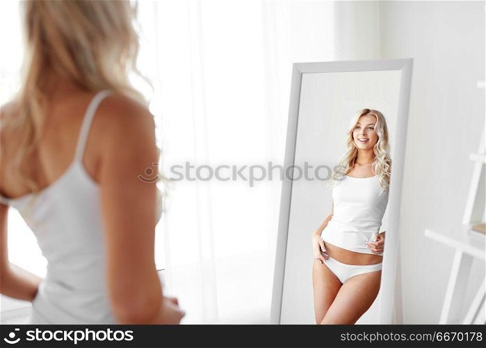 beauty, slimming and people concept - beautiful young woman in white underwear looking at mirror in morning at home. woman in underwear looking at mirror in morning. woman in underwear looking at mirror in morning