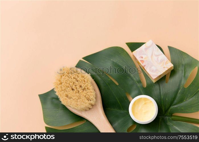beauty, spa and bath concept - close up of crafted soap bar, natural bristle wooden brush with body butter and monstera deliciosa leaf on beige background. natural soap, brush, body butter and monstera