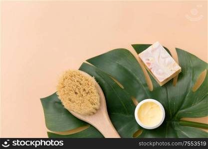 beauty, spa and bath concept - close up of crafted soap bar, natural bristle wooden brush with body butter and monstera deliciosa leaf on beige background. natural soap, brush, body butter and monstera