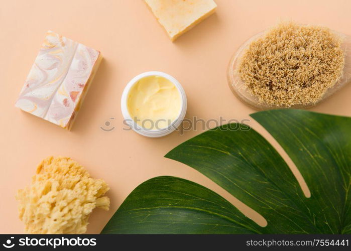 beauty, spa and bath concept - close up of crafted soap bar, natural bristle wooden brush, body butter with sponge and monstera deliciosa leaf on beige background. natural soap, brush, sponge and body butter