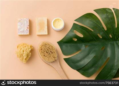 beauty, spa and bath concept - close up of crafted soap bar, natural bristle wooden brush, body butter with sponge and monstera deliciosa leaf on beige background. natural soap, brush, sponge and body butter