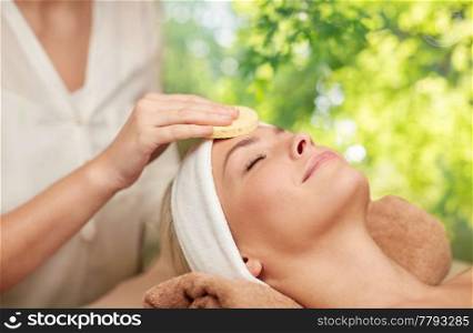 beauty, spa and wellness concept - close up of beautiful woman having face massage with sponge over green natural background. close up of woman having face massage in spa
