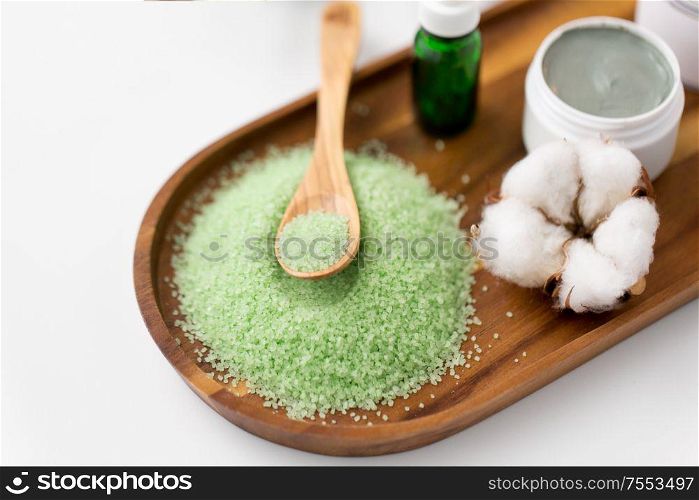 beauty, spa and wellness concept - green bath salt with wooden spoon, blue clay mask, cotton flower and serum on tray. bath salt, serum, clay mask and cotton on tray