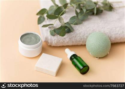 beauty, spa and wellness concept - serum or essential oil, clay mask, soap bar, konjac sponge and eucalyptus cinerea on bath towel. serum, clay mask, oil and eucalyptus on bath towel