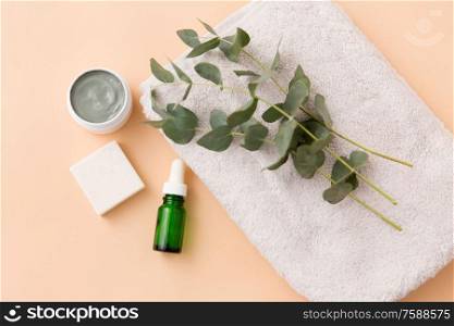 beauty, spa and wellness concept - serum or essential oil, clay mask, soap bar and eucalyptus cinerea on bath towel. serum, clay mask, oil and eucalyptus on bath towel