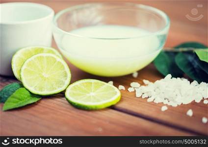 beauty, spa, body care, natural cosmetics and wellness concept - close up of citrus body lotion in glass bowl and sea salt with limes on wooden table. close up of body lotion in bowl and limes on wood