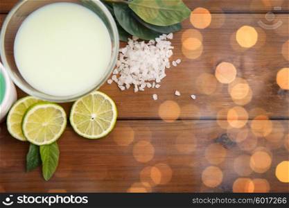 beauty, spa, body care, natural cosmetics and wellness concept - close up of citrus body lotion in glass bowl and sea salt with limes on wooden table