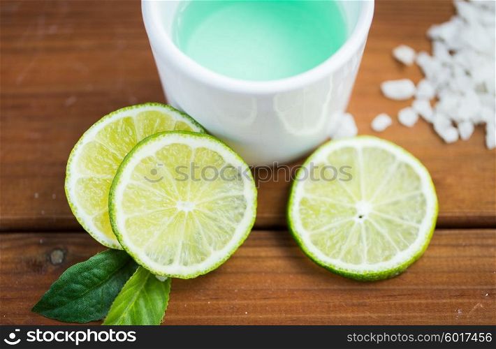 beauty, spa, body care, natural cosmetics and wellness concept - close up of citrus body lotion in cup and sea salt with limes on wooden table
