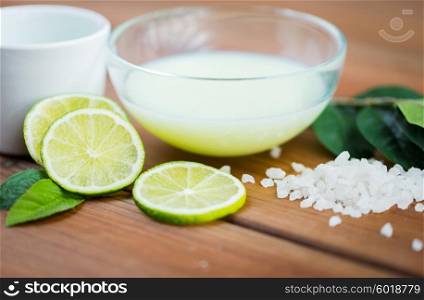 beauty, spa, body care, natural cosmetics and wellness concept - close up of citrus body lotion in glass bowl and sea salt with limes on wooden table