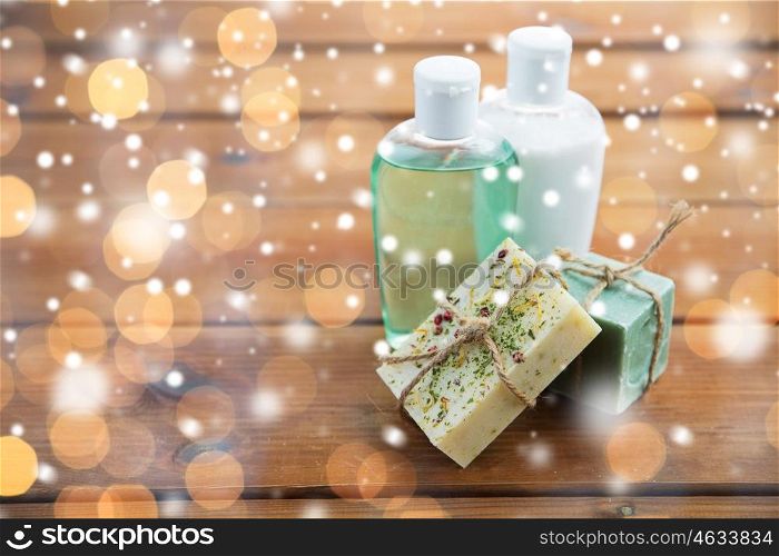 beauty, spa, bodycare, bath and natural cosmetics concept - handmade soap bars and lotion bottles on wooden table over lights and snow
