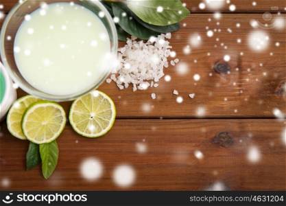 beauty, spa, bodycare, natural cosmetics and wellness concept - citrus body lotion in glass bowl and sea salt with limes on wooden table over snow