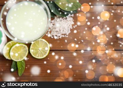 beauty, spa, bodycare, natural cosmetics and wellness concept - citrus body lotion in glass bowl and sea salt with limes on wooden table over lights and snow