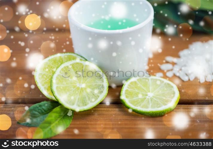 beauty, spa, bodycare, natural cosmetics and wellness concept - citrus body lotion in cup and sea salt with limes on wooden table over lights and snow