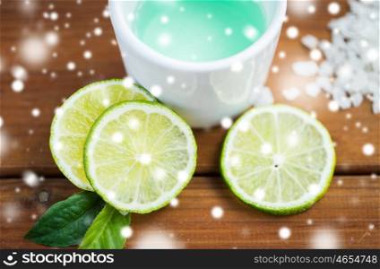 beauty, spa, bodycare, natural cosmetics and wellness concept - citrus body lotion in cup and sea salt with limes on wooden table over lights and snow
