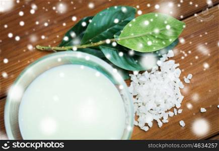 beauty, spa, bodycare, natural cosmetics and wellness concept - close up of body lotion in glass bowl and sea salt on wooden table over snow