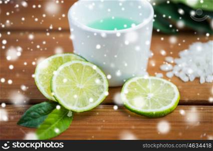 beauty, spa, bodycare, natural cosmetics and wellness concept - close up of citrus body lotion in cup and sea salt with limes on wooden table over snow