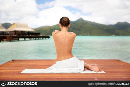 beauty, spa, luxury and bodycare concept - beautiful young woman in bath towel with bare top sitting on wooden pier over bungalow and island in french polynesia. beautiful young woman in white towel with bare top. beautiful young woman in white towel with bare top