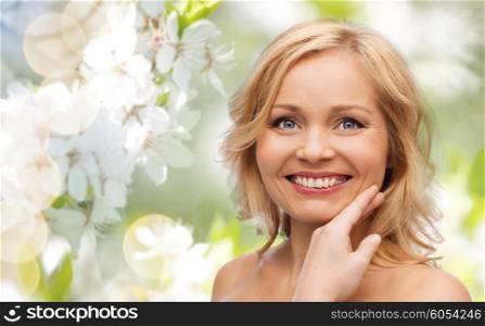 beauty, spring, skincare and natural cosmetics concept - smiling woman with bare shoulders touching face over cherry blossom background