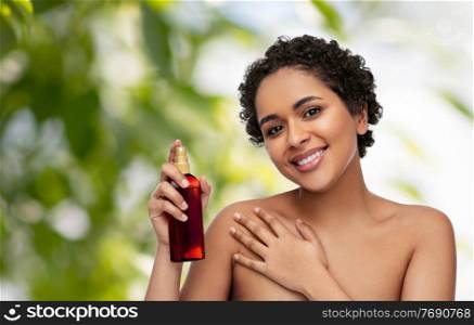beauty, sun protection and bodycare concept - portrait of happy smiling young african american woman sunscreen oil spray in bottle over green natural background. young african american woman with unscreen oil