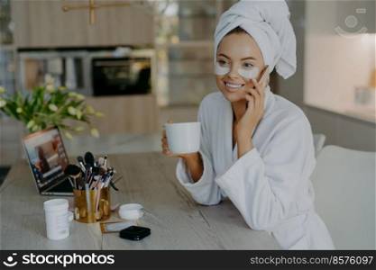 Beauty treatment personal care and hygiene concept. Lovely young woman applies cosmetic patches under eyes wears bathrobe drinks coffee talks to friend via smartphone sits at table over home interior