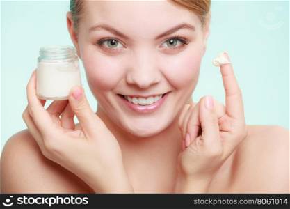 Beauty treatment. Woman applying moisturizing cream on face, holding jar with skin care product, studio shot on green blue background