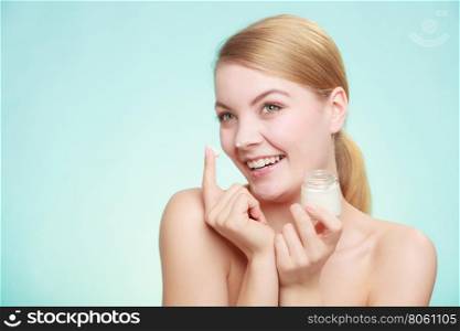Beauty treatment. Woman applying moisturizing cream on face, holding jar with skin care product, studio shot on green blue background