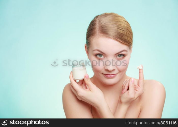 Beauty treatment. Woman applying moisturizing cream on face, holding jar with skin care product, studio shot on green blue background