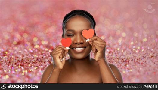 beauty, valentine&rsquo;s day and luxury concept - portrait of happy smiling young african american woman with bare shoulders with red hearts covering her eye over pink glitter background. smiling african american woman with red hearts