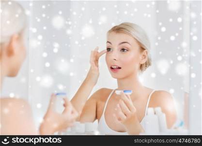 beauty, vision, eyesight, ophthalmology and people concept - young woman putting on contact lenses at mirror in home bathroom over snow