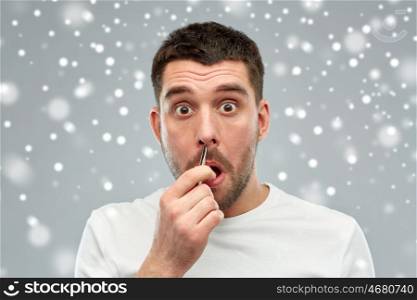 beauty, winter, christmas and people concept - smiling young man with tweezers tweezing hair frome nose over snow on gray background