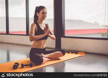 Beauty woman doing yoga and raise hand or pay obeisance in fitness workouts training gym center. Lifestyle sport woman sitting on mat with sport equipment and exercise dumbbells background