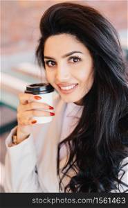 Beauty, youth, lifestyle and relaxation concept. Young brunette female with well-shaped thin eyebrows, healthy skin and red manicure holding takeaway coffee looking pleasantly into camera smiling