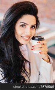 Beauty, youth, lifestyle and relaxation concept. Young brunette female with well-shaped thin eyebrows, healthy skin and red manicure holding takeaway coffee looking pleasantly into camera smiling